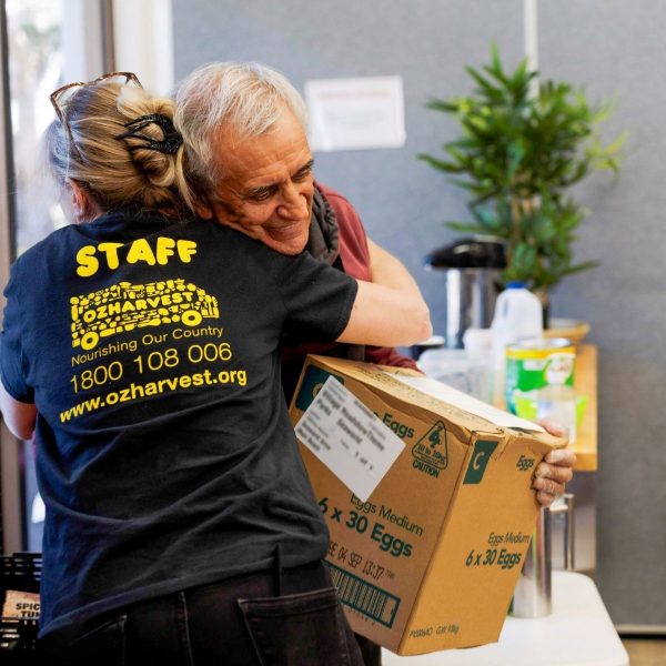 An OzHarvest staff member in a branded shirt hugs a man holding a carton of eggs