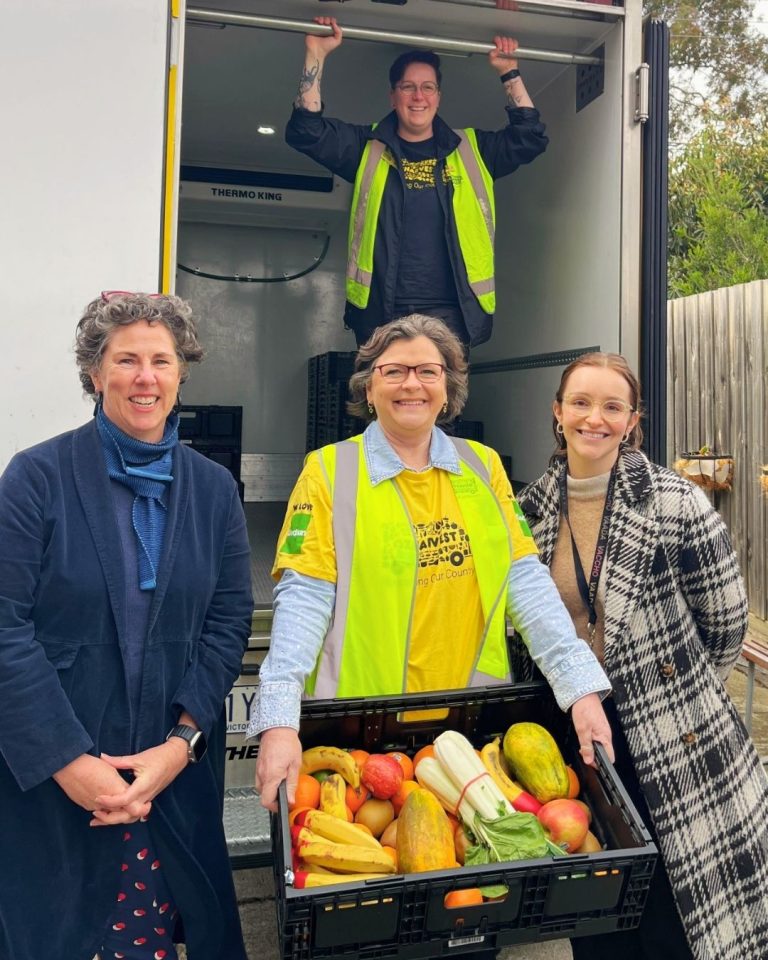 Four people by a refrigerated OzHarvest truck, a woman in a hi-vis vest holding a crate of rescued fruit and vegetables