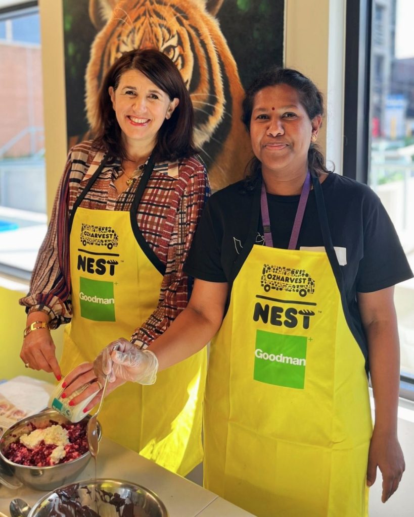Two women in OzHarvest NEST Goodman aprons prepare food together at a bench by a tiger mural
