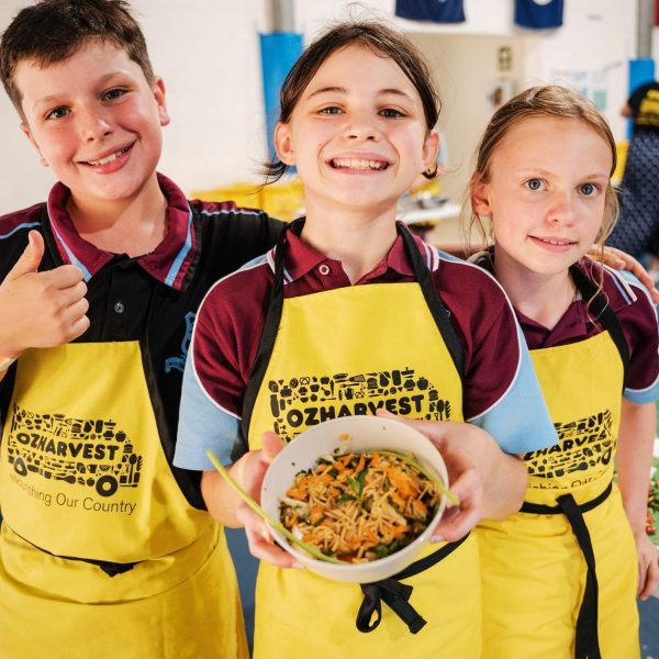 Three smiling school children in yellow OzHarvest aprons holding a bowl of fresh stir-fry they cooked