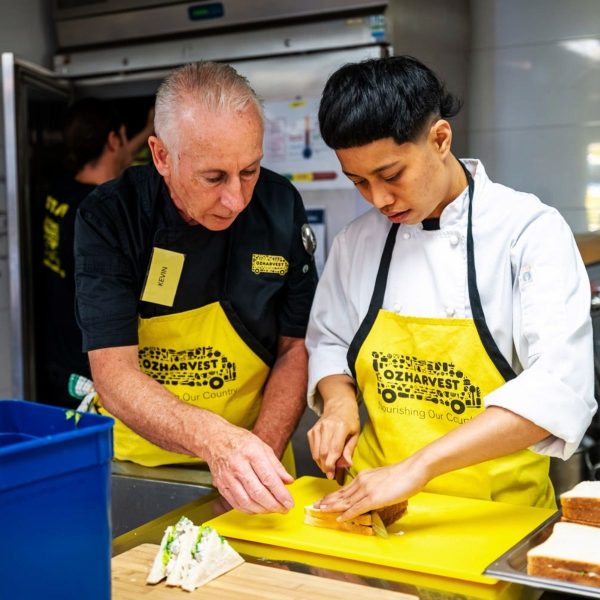 OzHarvest volunteer in apron showing a young chef how to slice sandwiches in a commercial kitchen