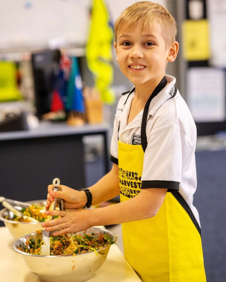 Smiling schoolboy in a yellow OzHarvest apron uses tongs to toss a fresh salad in a metal bowl in a FEAST class
