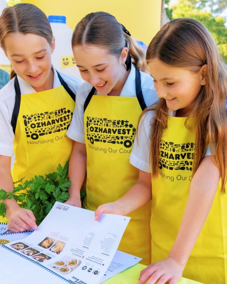 Three schoolgirls in yellow OzHarvest aprons read a recipe booklet together outdoors