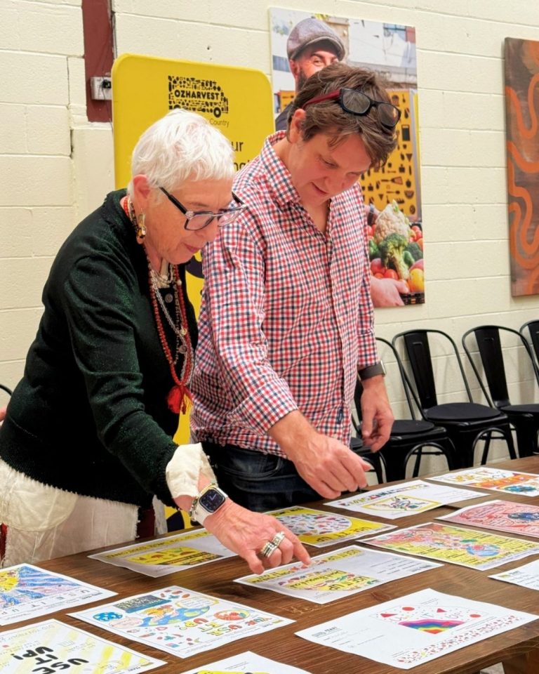 Two people arrange 'Use It Up' artwork posters on a table beneath an OzHarvest banner at a workshop