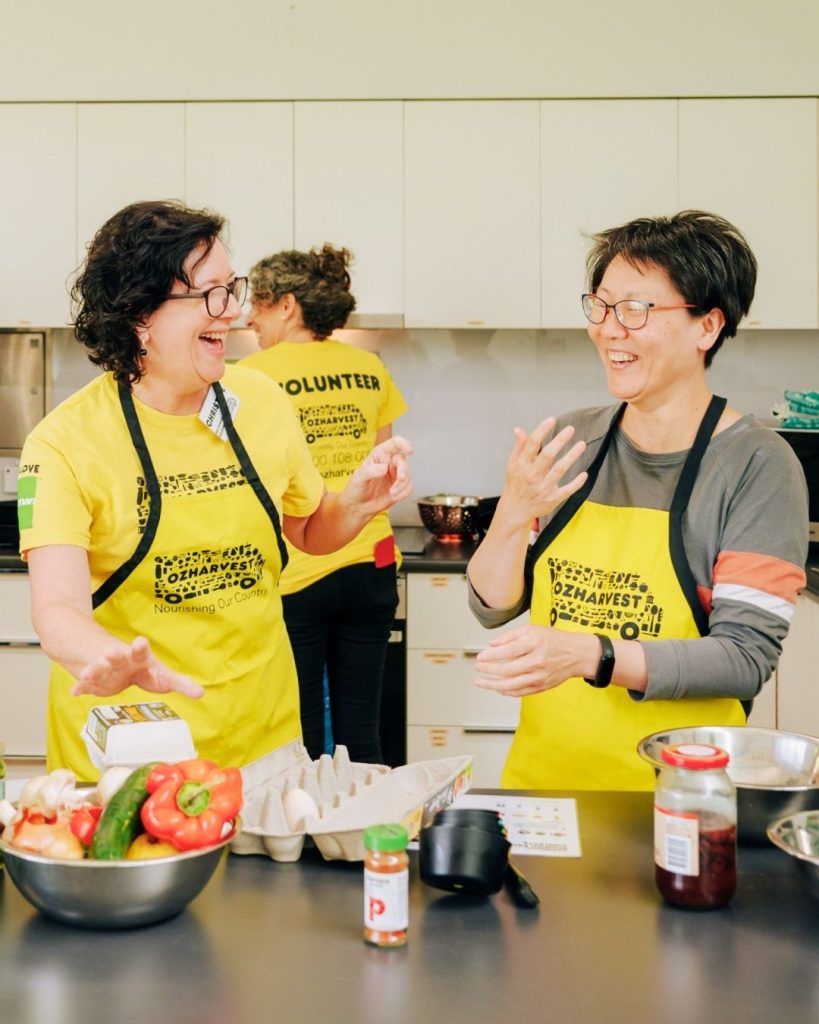 Two women in yellow OzHarvest aprons laugh together while cooking with fresh vegetables in a kitchen