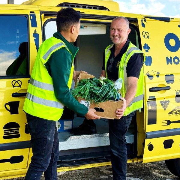 Two men in hi-vis vests carry a box of fresh greens beside a bright yellow OzHarvest van