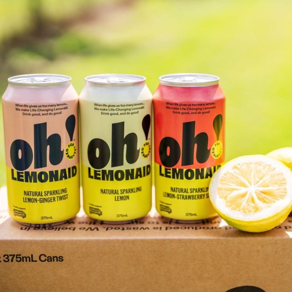 Three cans of Oh! Lemonaid natural sparkling lemonade beside a halved lemon on a cardboard box