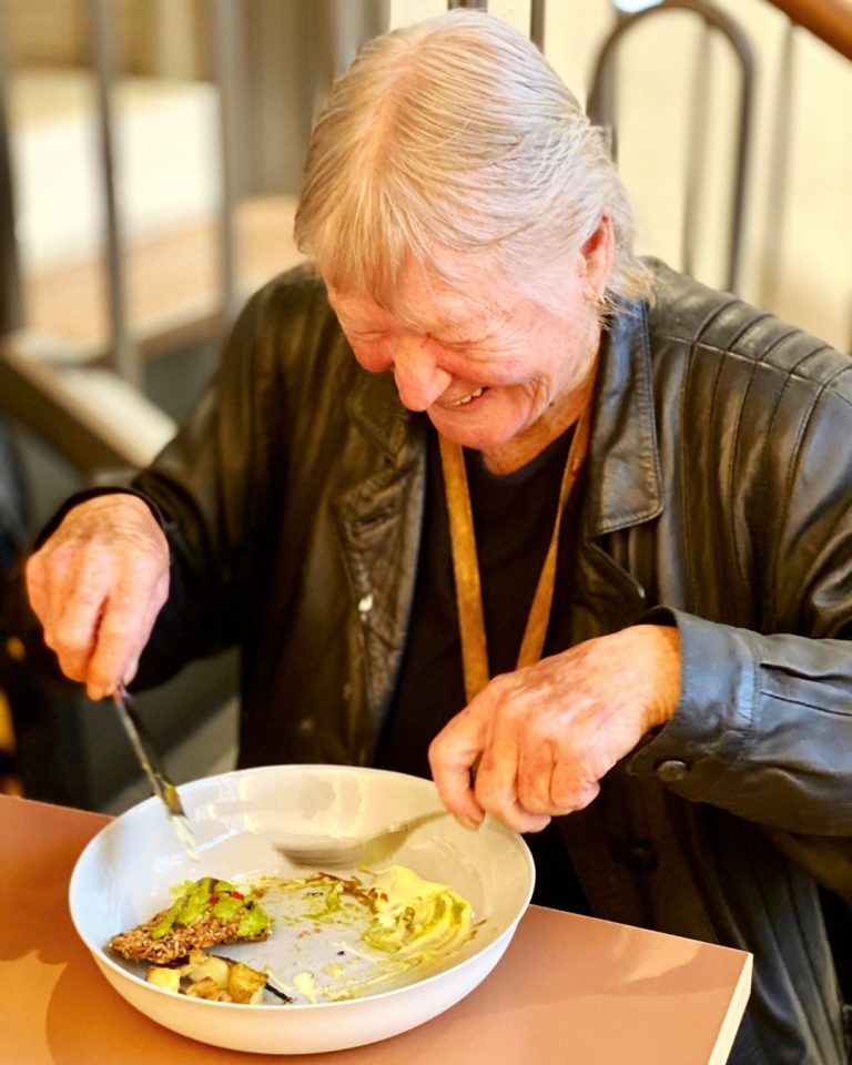 Older person in leather jacket smiling while eating a plated meal at a Refettorio dining table