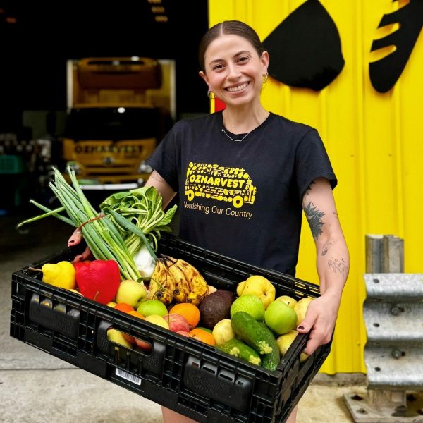 Smiling woman in OzHarvest t-shirt holds a black crate brimming with rescued fruit and vegetables by a yellow truck