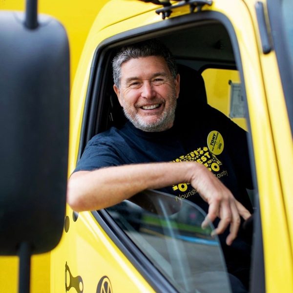 Smiling grey-bearded man leans out the window of a yellow OzHarvest truck cab