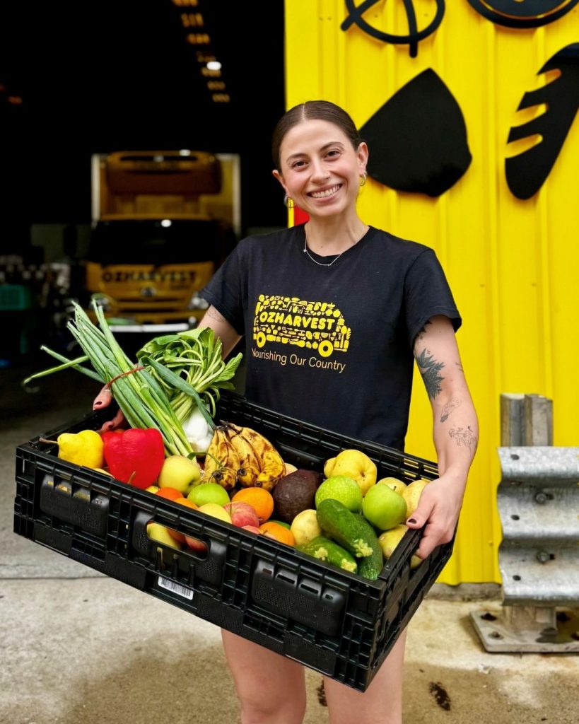 Smiling OzHarvest worker holding a crate of mixed fruit and vegetables by a yellow truck