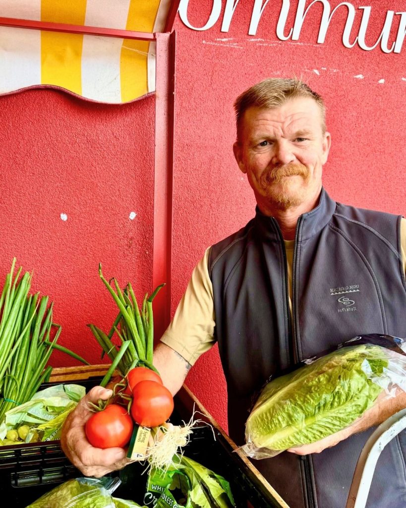 Man in vest holding tomatoes and lettuce at a community market stall with fresh produce