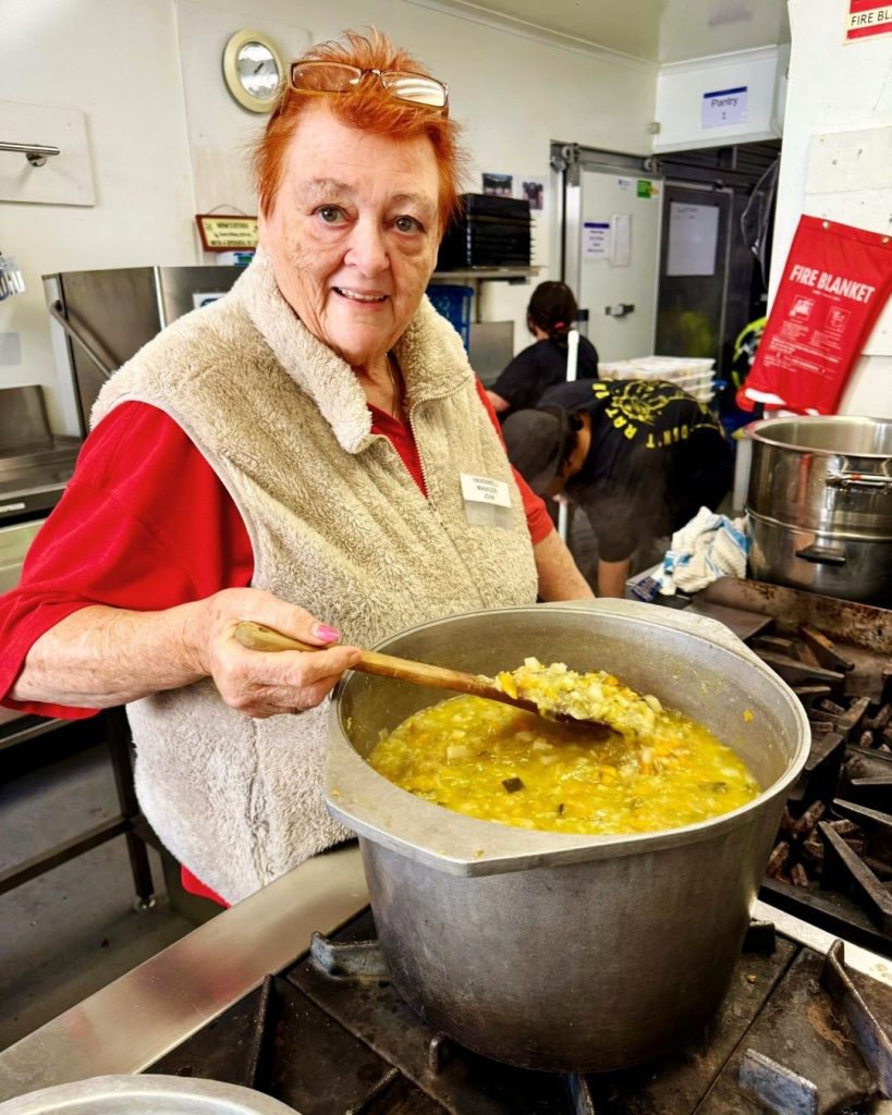 Smiling OzHarvest volunteer stirring a large pot of soup with a wooden spoon in a community kitchen