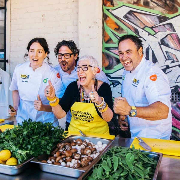 Chefs and a woman in an OzHarvest apron laugh together at an outdoor cooking station with fresh produce