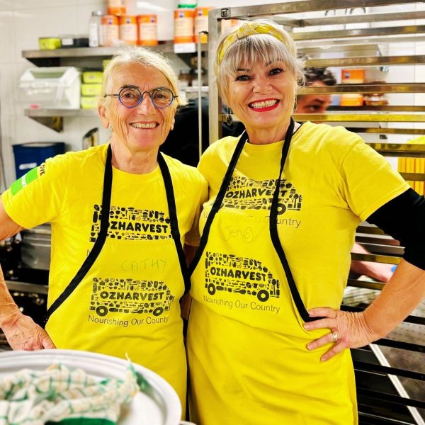 Two smiling women in yellow OzHarvest shirts and black aprons stand together in a commercial kitchen