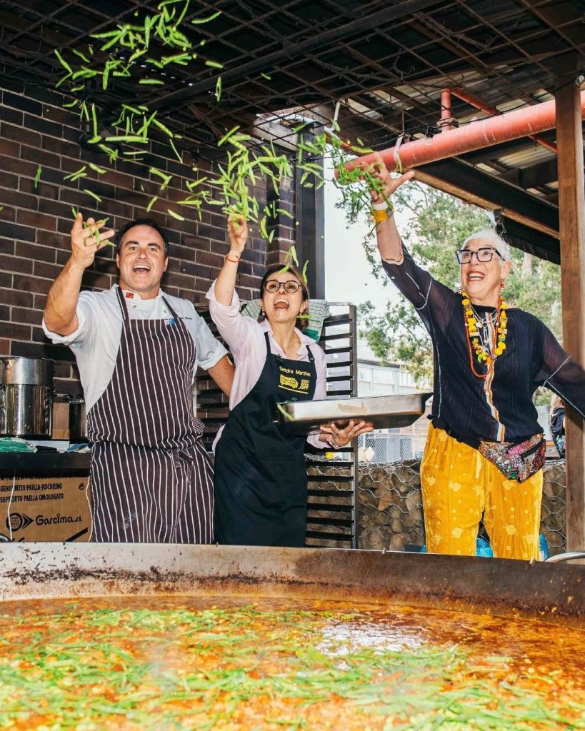 Chefs and volunteers tossing green beans into the air above a giant paella pan at an OzHarvest event