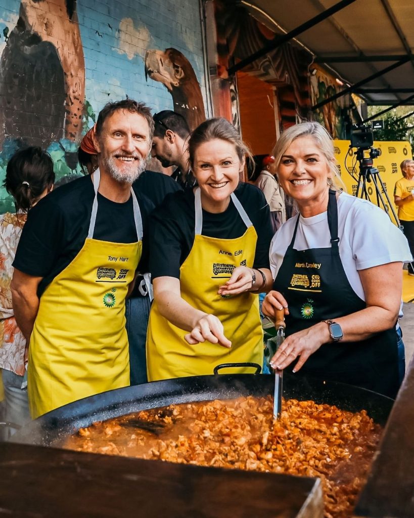 Volunteers in yellow BP aprons laughing while cooking a huge paella at an OzHarvest community feast