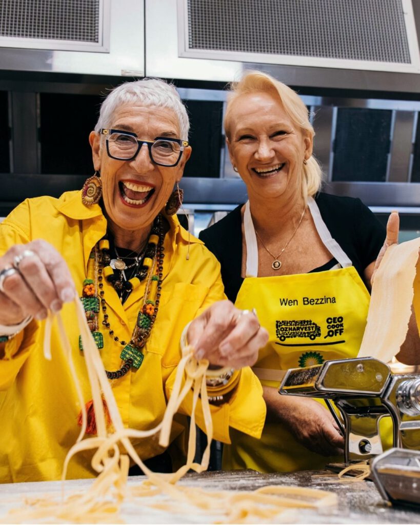 Two women laughing while making fresh pasta on a machine, one in OzHarvest CEO CookOff apron